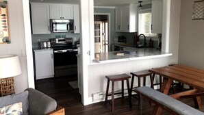 Cozinha compacta com armários brancos, micro-ondas e torradeira. Há uma mesa de jantar de madeira com bancos e uma luminária.