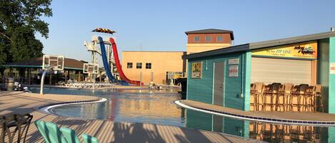 A pool area with a water slide, lounge chairs, and a building with a 'Buy Your ZIP Line Tickets Here!' sign.
