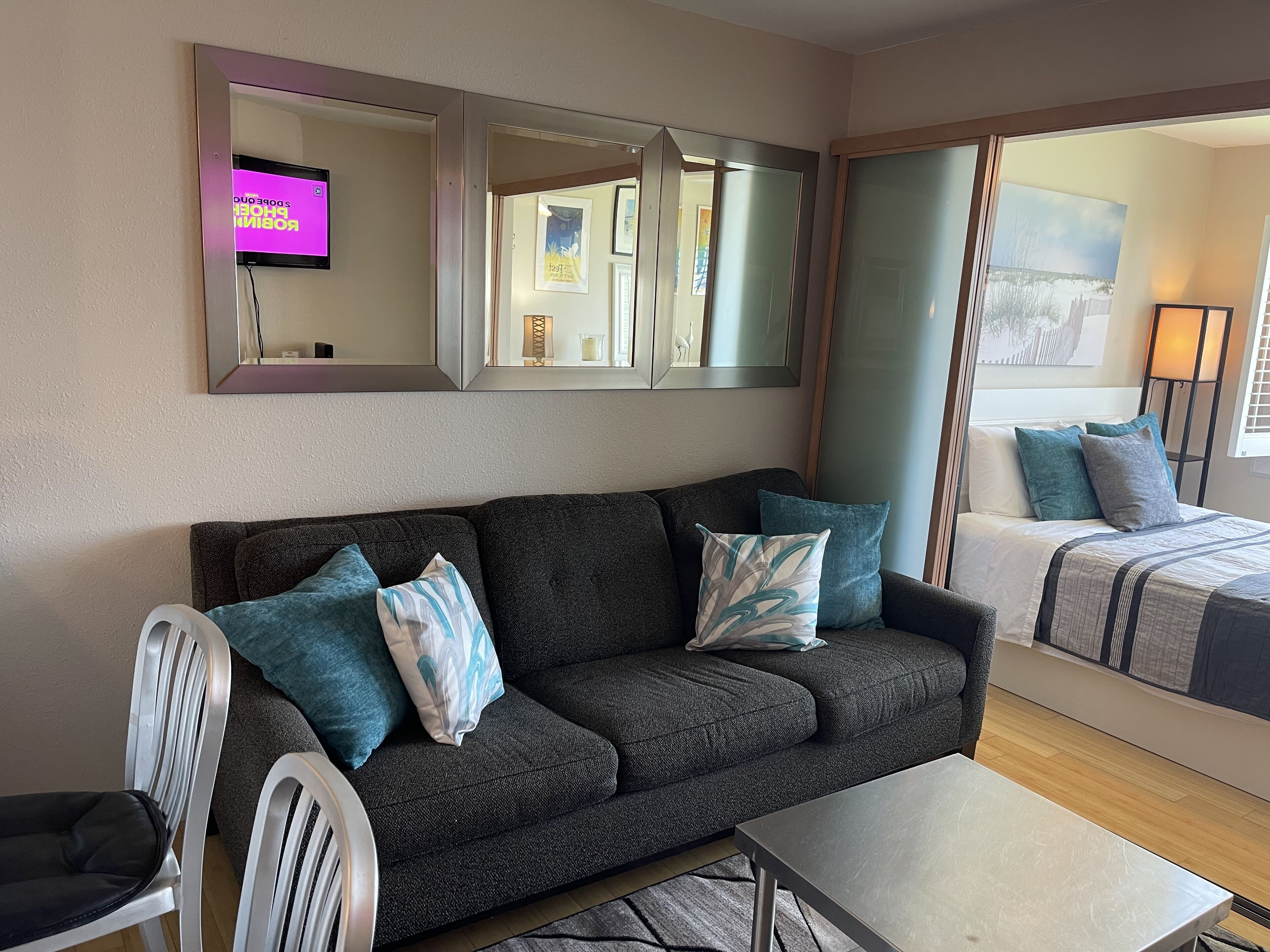 Un salón moderno con un sofá gris oscuro, una silla blanca y una mesita. Hay dos camas con ropa de cama azul y gris. Un espejo grande y un cuadro están colgados en la pared.