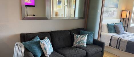 Un salón moderno con un sofá gris oscuro, una silla blanca y una mesita. Hay dos camas con ropa de cama azul y gris. Un espejo grande y un cuadro están colgados en la pared.