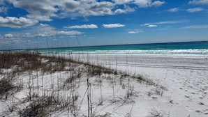 Ein Strand mit weißem Sand und vereinzelter Vegetation unter einem teils bewölkten Himmel.