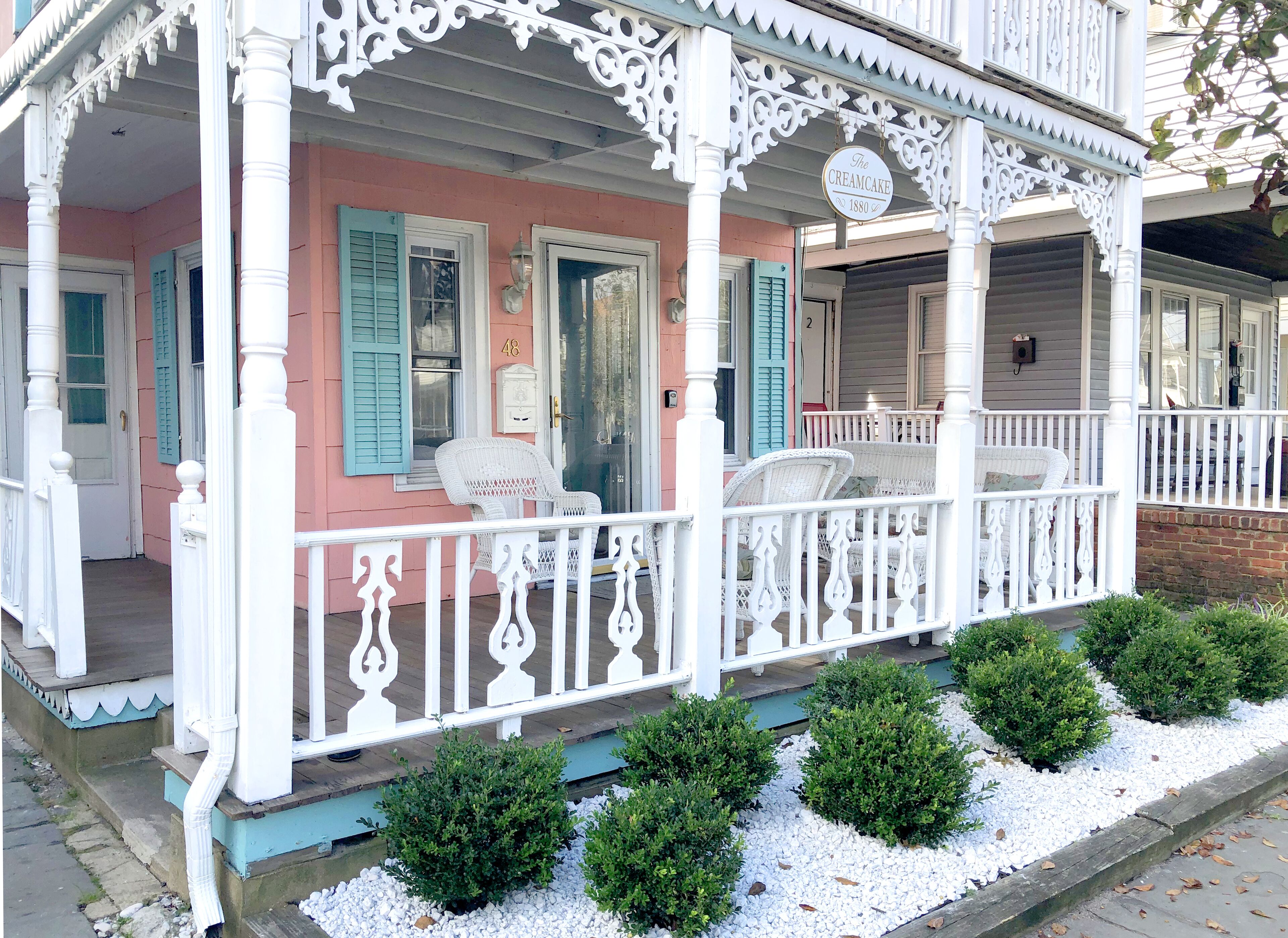 A pink building with white porch railings and blue shutters, featuring a white chair and a small white table.