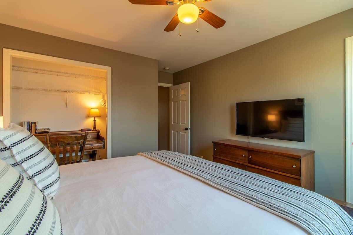 Quarto com cama, televisão, cómoda de madeira, escrivaninha com candeeiro e ventoinha no teto.