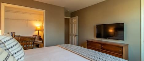 Quarto com cama, televisão, cómoda de madeira, escrivaninha com candeeiro e ventoinha no teto.