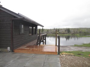 Una cabaña de madera con una terraza de madera que conduce a un muelle sobre un lago tranquilo, rodeada de árboles.