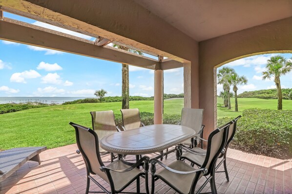 A covered patio with a round table and chairs, overlooking a golf course and the ocean. - Stunning First-Floor Turtle Dunes Villa! Just Steps from Pool and Ocean! (Fernandina Beach)