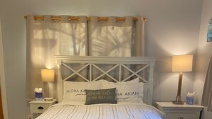 A neatly made bed with a white headboard, a bedside table with a lamp, and a wall with a sign. - HALE NIHI, Paia, Maui (Paia)