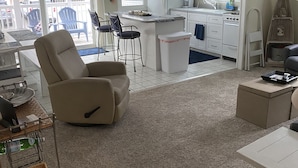 Un soggiorno spazioso con una poltrona beige, un tavolo da pranzo con sedie, una cucina con mobili bianchi e vista all'esterno attraverso ampie finestre.