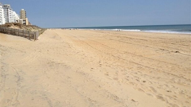 Una spiaggia ampia e sabbiosa con una passerella di legno sulla sinistra, che conduce a una fila di edifici.