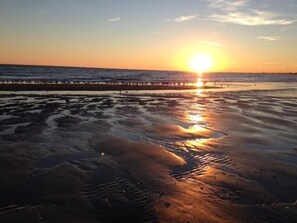 Ein Strand bei Sonnenuntergang, die Sonne tief am Horizont, spiegelt sich im feuchten Sand.