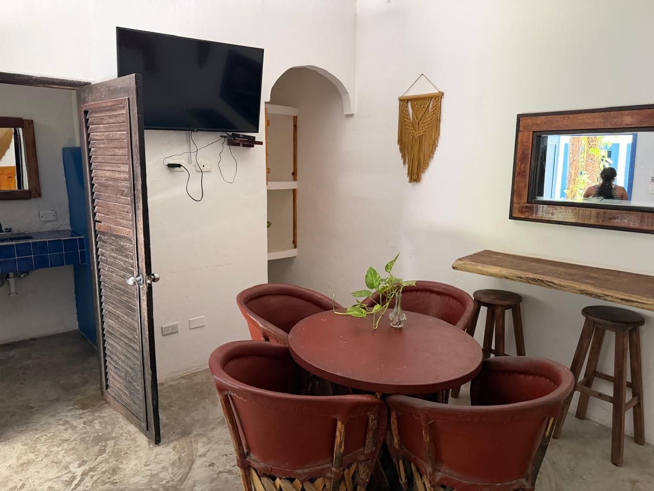 A dining area with a round table and chairs, a television mounted on the wall, and a kitchen area with blue tiles.