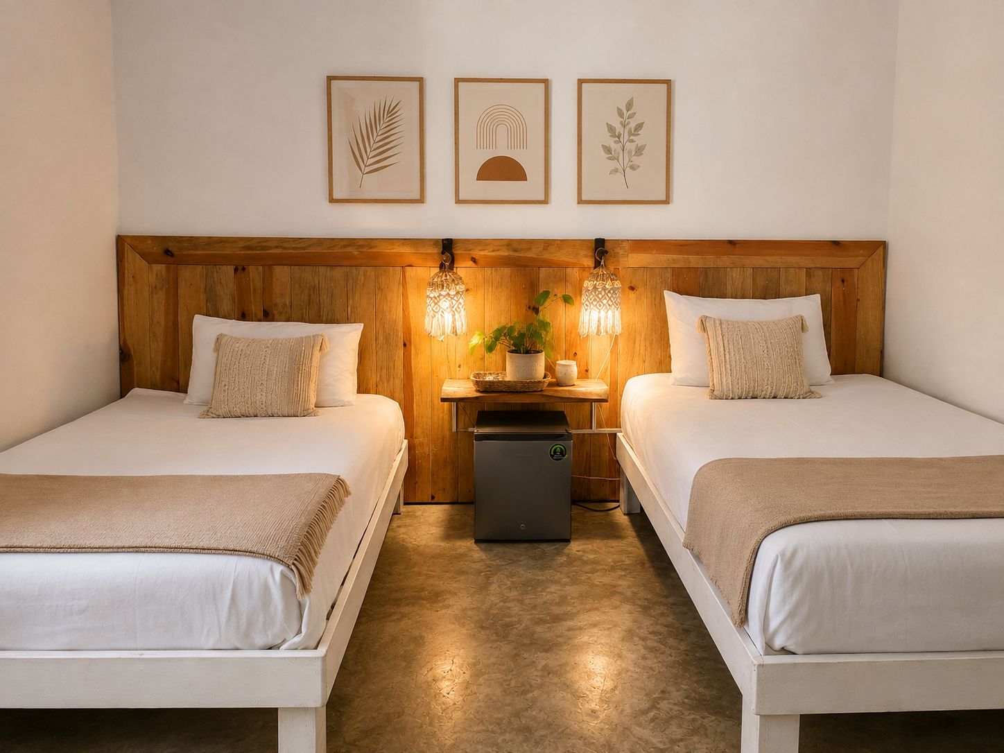 A hotel room with two beds, a wooden headboard, a small table with a lamp, and framed artwork on the wall.