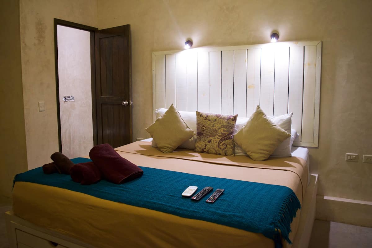 A hotel room with a bed, pillows, a blue blanket, and remote controls.