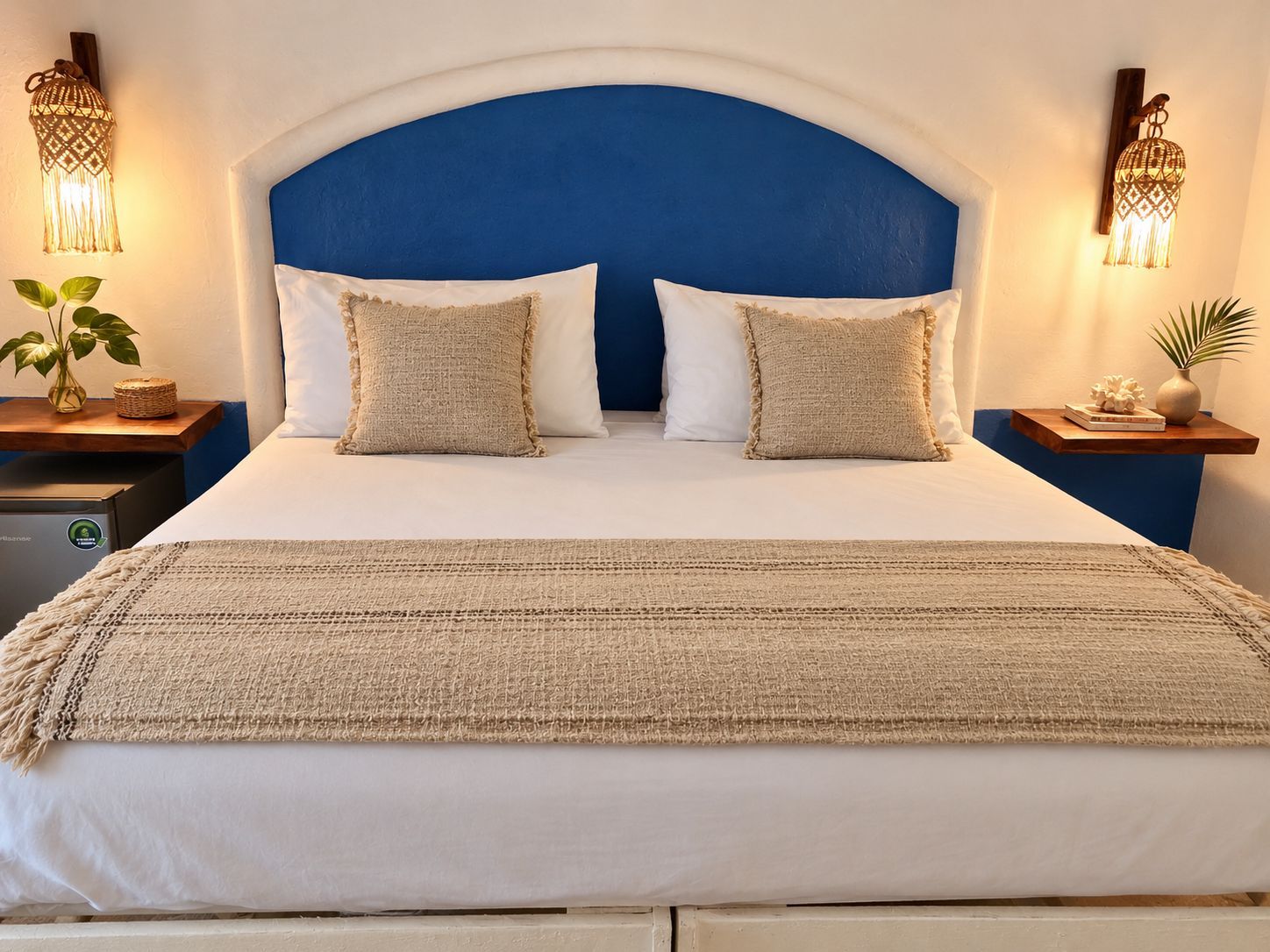 Un lit avec une tête de lit bleue, deux oreillers blancs et un plaid beige. Il y a deux tables de chevet, chacune surmontée d’une lampe décorative et d’une petite plante.