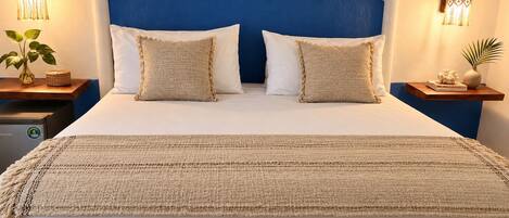 Un lit avec une tête de lit bleue, deux oreillers blancs et un plaid beige. Il y a deux tables de chevet, chacune surmontée d’une lampe décorative et d’une petite plante.