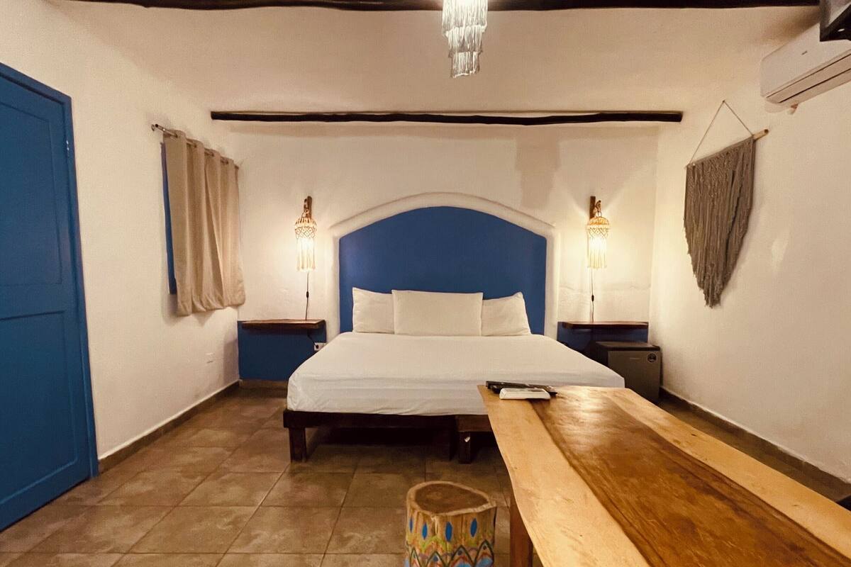 A bedroom with a bed, bedside tables, a bench, a colorful trash bin, and a chandelier.