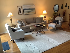 Un salon comprenant un canapé, deux fauteuils, une table basse et deux lampes. Un tableau encadré est accroché au mur, et un miroir se trouve au-dessus de la table basse.
