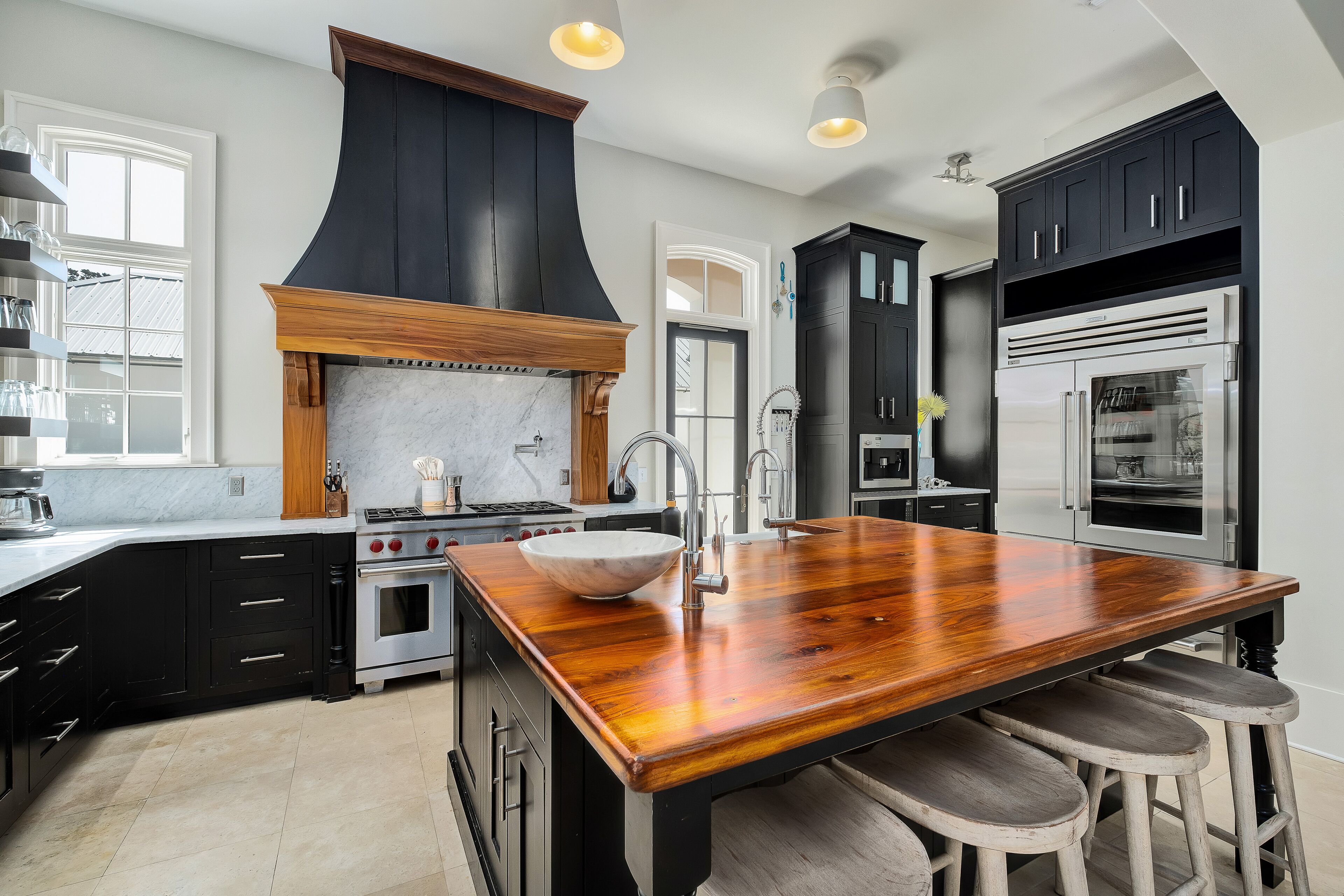 A modern kitchen with dark cabinetry, a large island with stools, a built-in oven, and a stainless steel refrigerator.