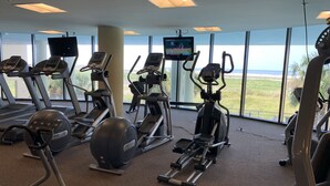 Ein Fitnessraum mit Laufbändern, Crosstrainern und einem Ergometer, mit großen Fenstern und Blick auf den Strand.