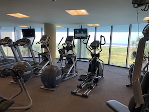 Une salle de sport équipée de tapis de course, de vélos elliptiques et d’un vélo d’appartement, avec de grandes fenêtres donnant sur la plage.