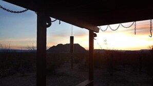 Ein überdachter Außenbereich mit einer Bank, Ketten, die von der Decke hängen, und Blick auf einen Sonnenuntergang über einer hügeligen Landschaft.