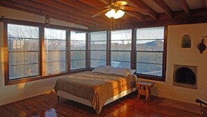 Ein Schlafzimmer mit Bett, Holzboden, großen Fenstern, Deckenventilator und Kamin.