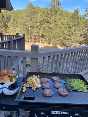 A grill with burgers, potatoes, and asparagus on a deck with a view of trees.