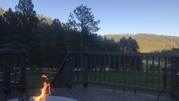 Una terraza con un hoyo para el fuego, rodeada por barandas de madera y vista a un campo cubierto de hierba y árboles.