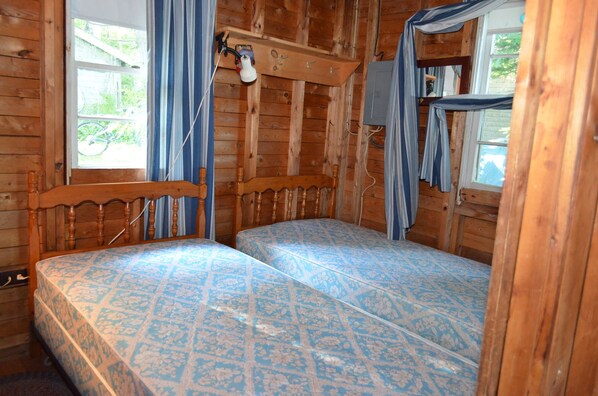 Una habitación tipo cabaña de madera con dos camas, una ventana con cortinas y un estante pequeño con una lámpara.