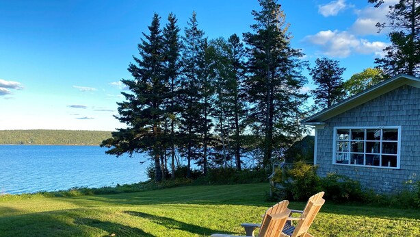 En ejendom ved søen med en træterrasse, to Adirondack-stole og en bygning i sten med et vindue.