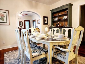 A dining room with a round table set for four, surrounded by white chairs with patterned upholstery. A green bookcase with various items is visible in the background. - 1940s Vintage Home meets comfortable beach retreat (Galveston)