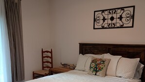 Une chambre à coucher comprenant un lit, une tête de lit en bois, un élément décoratif mural, une chaise et une table sur laquelle est posée une maquette de maison.