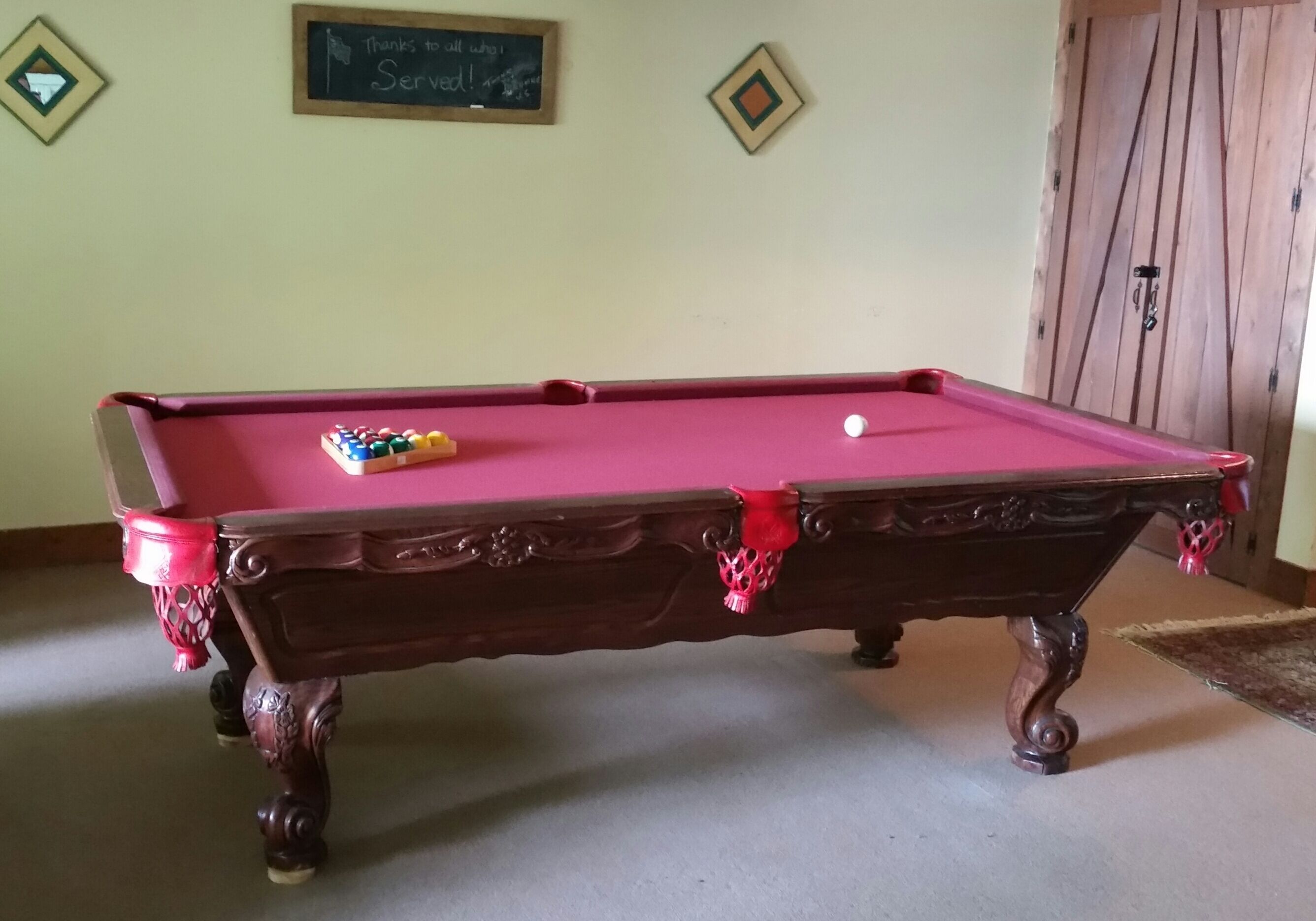 A pool table with a red felt surface, surrounded by a wooden frame with intricate carvings.