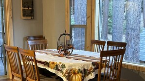 Une salle à manger avec une table et des chaises en bois, un chemin de table orné de motifs de citrouilles et une fenêtre donnant sur des arbres.