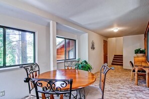 A dining area with a round wooden table and metal chairs, large windows with a view of trees, and a staircase leading to another level.
