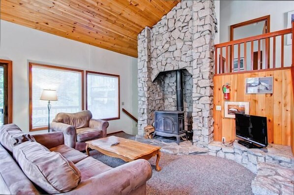 A cozy living room with a stone fireplace, wooden ceiling, and a TV on a wooden stand.