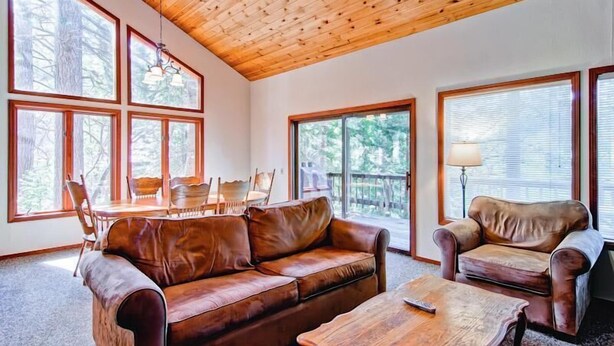 A cozy living room with a wooden ceiling, large windows, a leather sofa, a coffee table, and a chair.