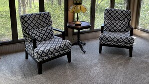 A room with two patterned armchairs, a small table with a lamp, and large windows offering a view of trees.