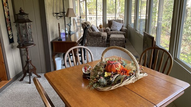 A cozy room with a wooden table, chairs, a wicker basket with autumn leaves, a lamp, and a view of trees through large windows.