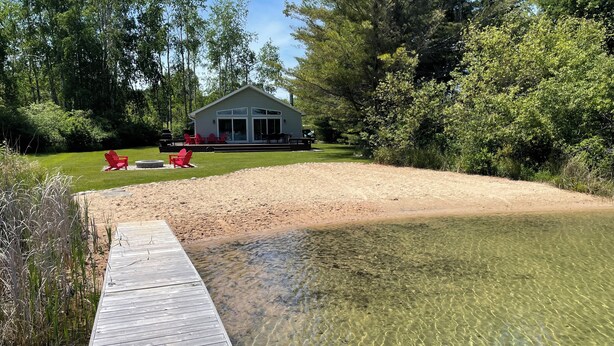 A wooden deck leading to a clear water body, surrounded by a grassy area with red chairs and a building in the background. - Hidden Springs on Long Lake  -  Sleeping Bear Dunes National Lakeshore (Honor)
