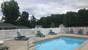 A rectangular pool with a metal ladder, surrounded by a white fence. There are several green lounge chairs and a small white table near the pool. - Hillside Motel (Lake George)
