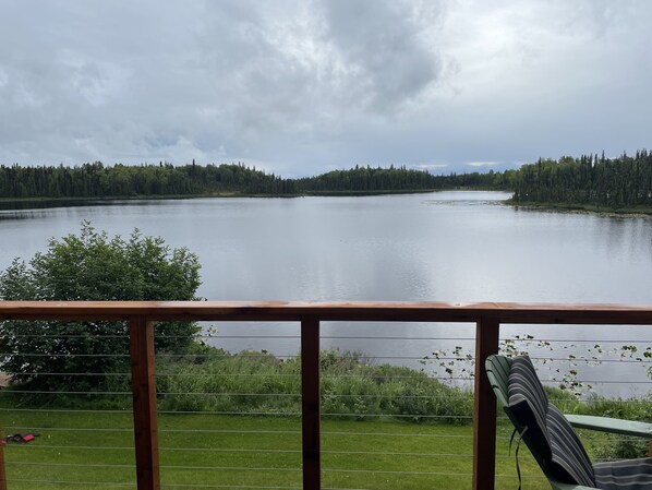 A lakeside view from a deck with a chair, a grassy area, and dense forest in the background. - Beautiful Lakefront Property Minutes From The Kenai River And Soldotna (Soldotna)