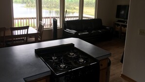 A kitchen with a stove, dining table, and chairs. There is a large window with a view of a lake and trees. A black leather couch is in the living area. - Beautiful Lakefront Property Minutes From The Kenai River And Soldotna (Soldotna)