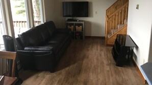 A living room with a black leather sofa, a television on a stand, a wooden staircase, and a view of the outdoors through large windows. - Beautiful Lakefront Property Minutes From The Kenai River And Soldotna (Soldotna)