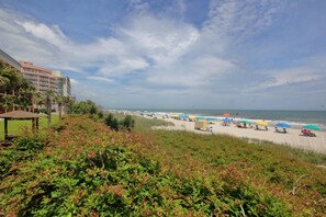 A beach with a hotel in the background, a sandy shore, and people relaxing under umbrellas. - Myrtle Beach Oceanfront Condo with Huge Sundeck Balcony (Myrtle Beach)