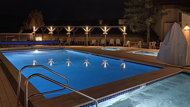 Une piscine nocturne dotée d’une terrasse en bois, de chaises longues et d’un espace couvert éclairé.
