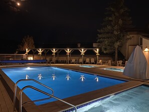 Una piscina nocturna con cubierta de madera, sillones de descanso y una zona techada con luces.