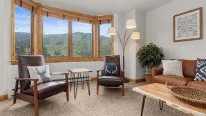 Un salon chaleureux avec une grande fenêtre, un canapé en cuir marron, une table basse en bois et deux fauteuils en cuir agrémentés de coussins décoratifs.