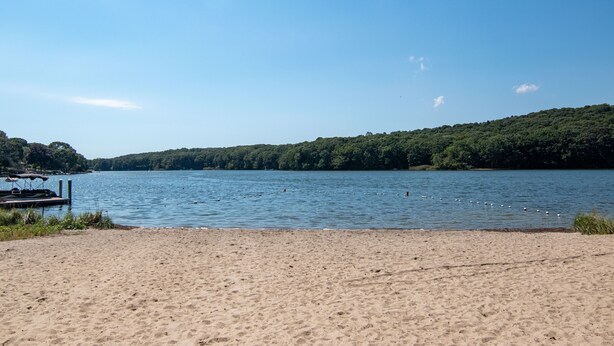 A beach with a dock and a forested hill in the background. - Cozy Escape in Pettaquamscutt Neighborhood - Your Ideal Narragansett Getaway (Narragansett)