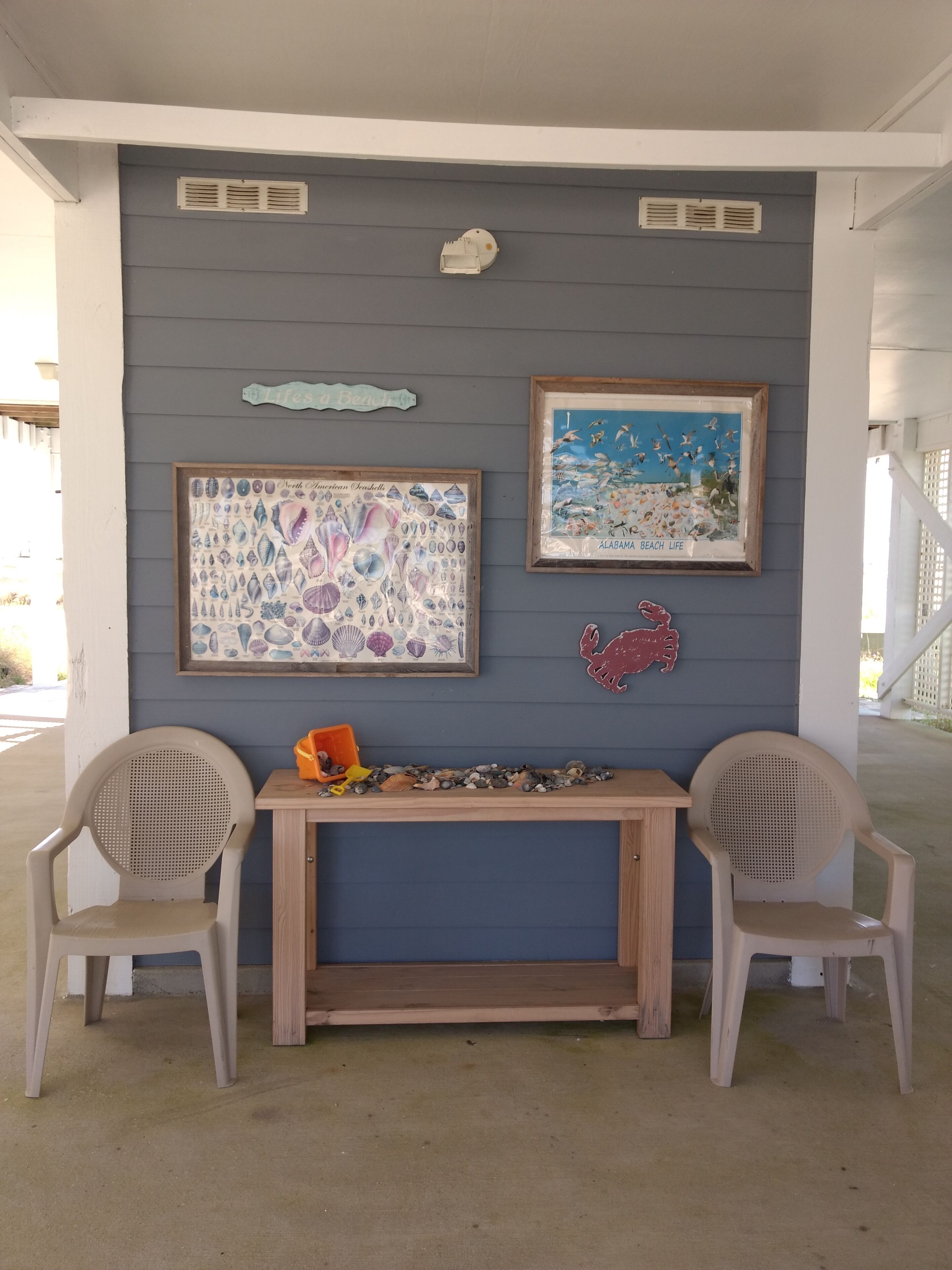 A covered outdoor seating area with two chairs, a table, and wall decorations including a framed seascape and a framed shell collection.